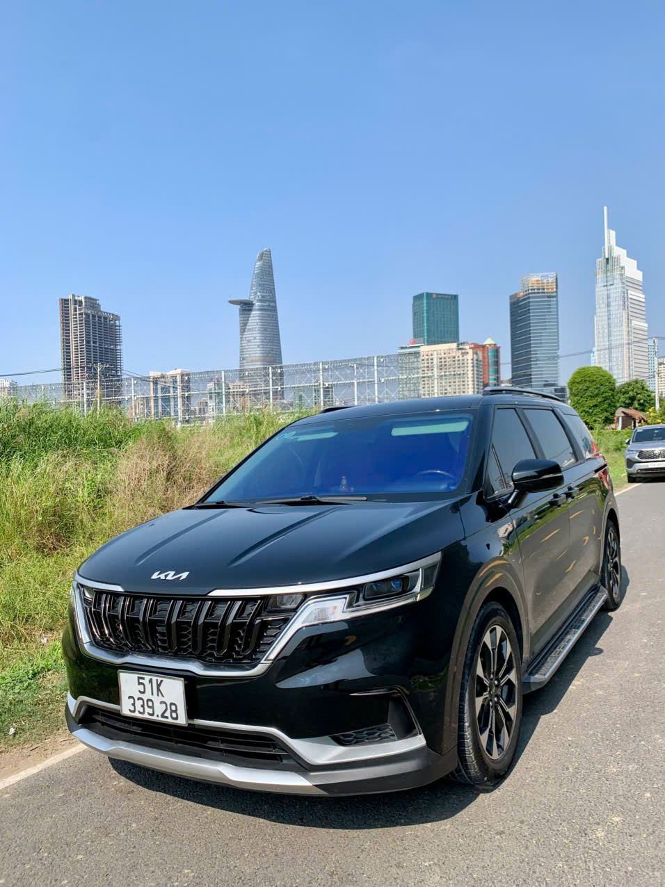 Xe Kia Carnival VIP góc 3/4 trước với skyline thành phố, dịch vụ đưa đón sân bay